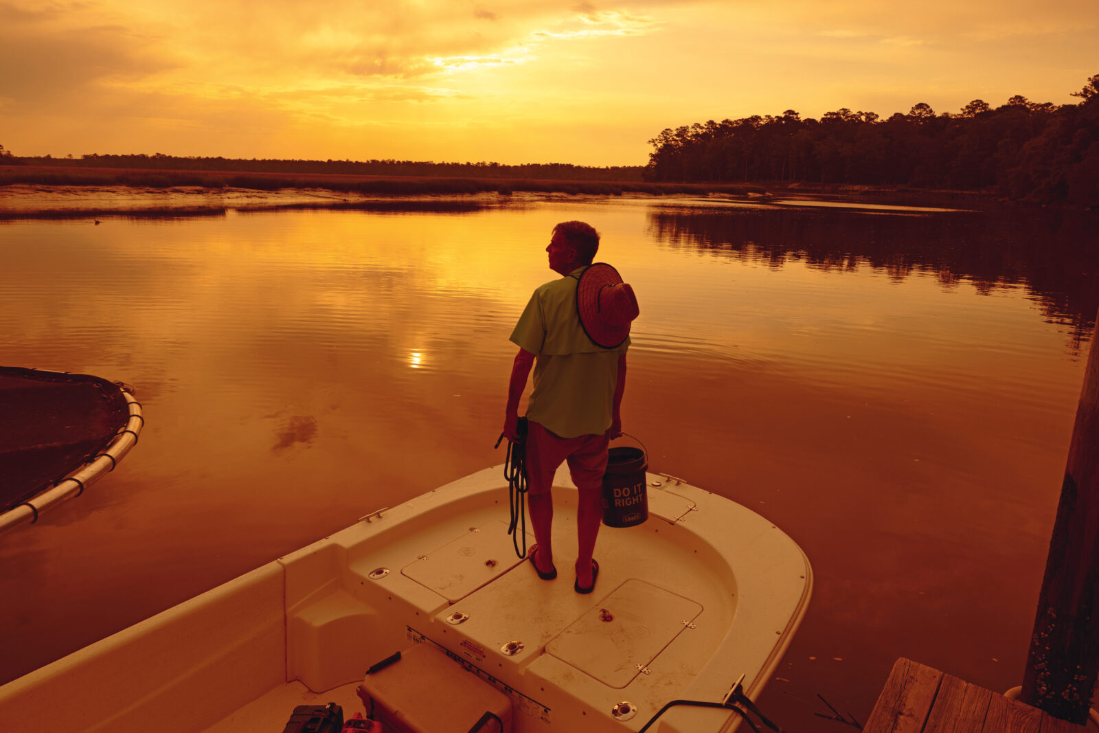 A man stands on the front of a boat at a red sunrise