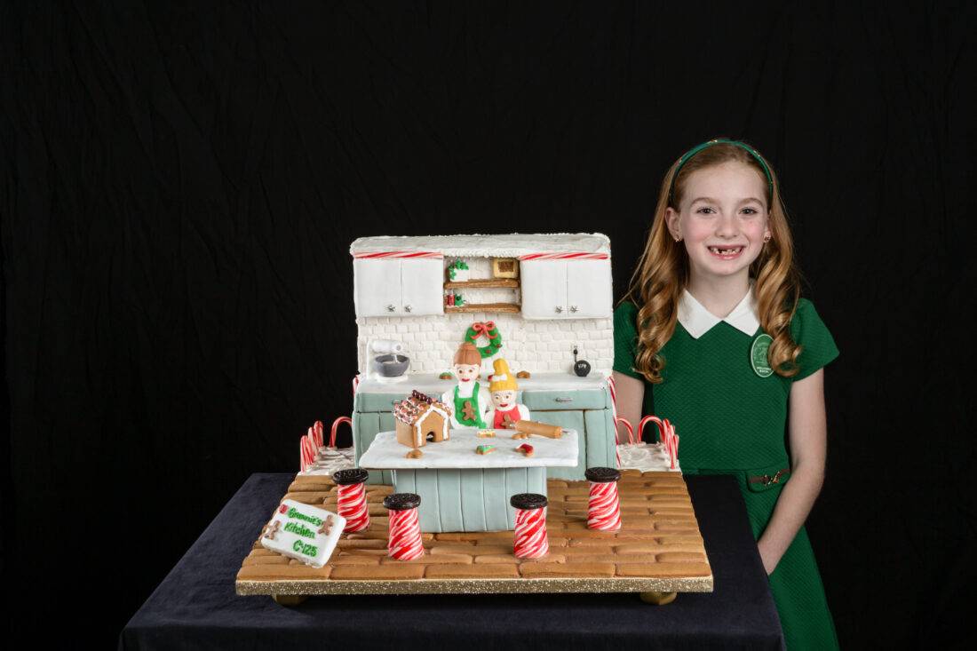 A girl stands with a gingerbread house of a kitchen