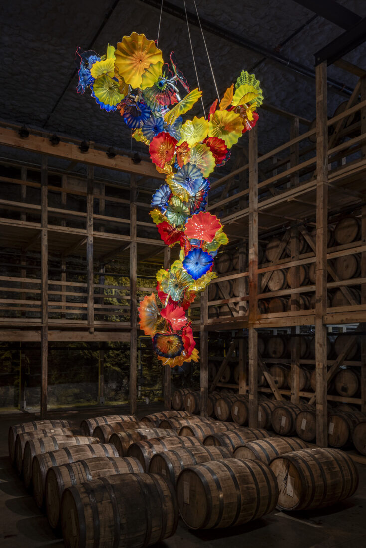 A rainbow glass sculpture hangs above bourbon barrels
