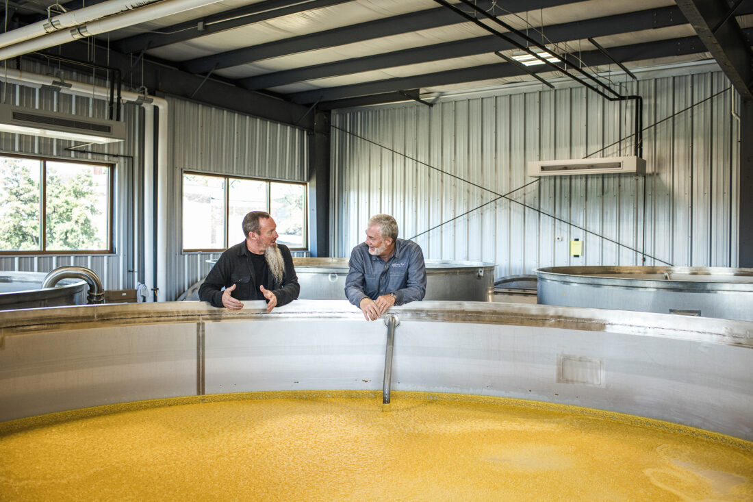 Two men stand over a distilling tank