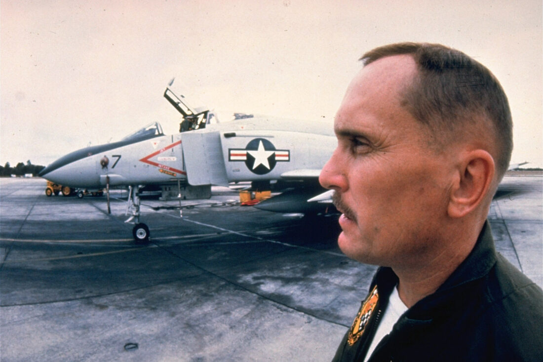 A profile of a man on a movie set with a military jet behind him.