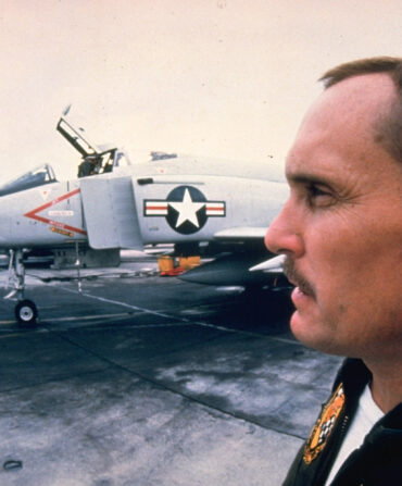 A profile of a man on a movie set with a military jet behind him.