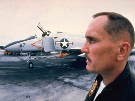 A profile of a man on a movie set with a military jet behind him.
