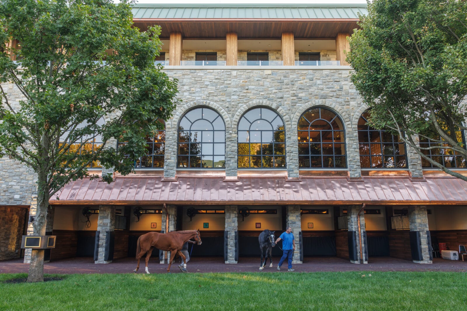 Two horses led by people outside of a paddock building