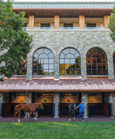 Two horses led by people outside of a paddock building