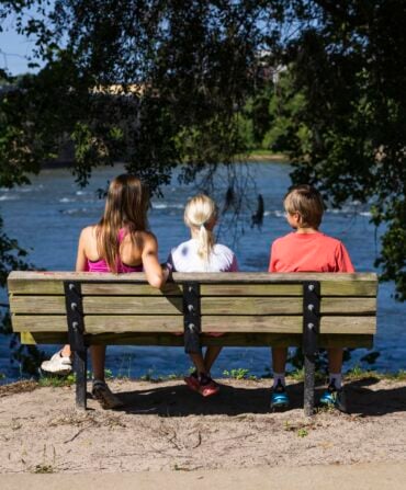 A woman and two children on a park bench facing a river