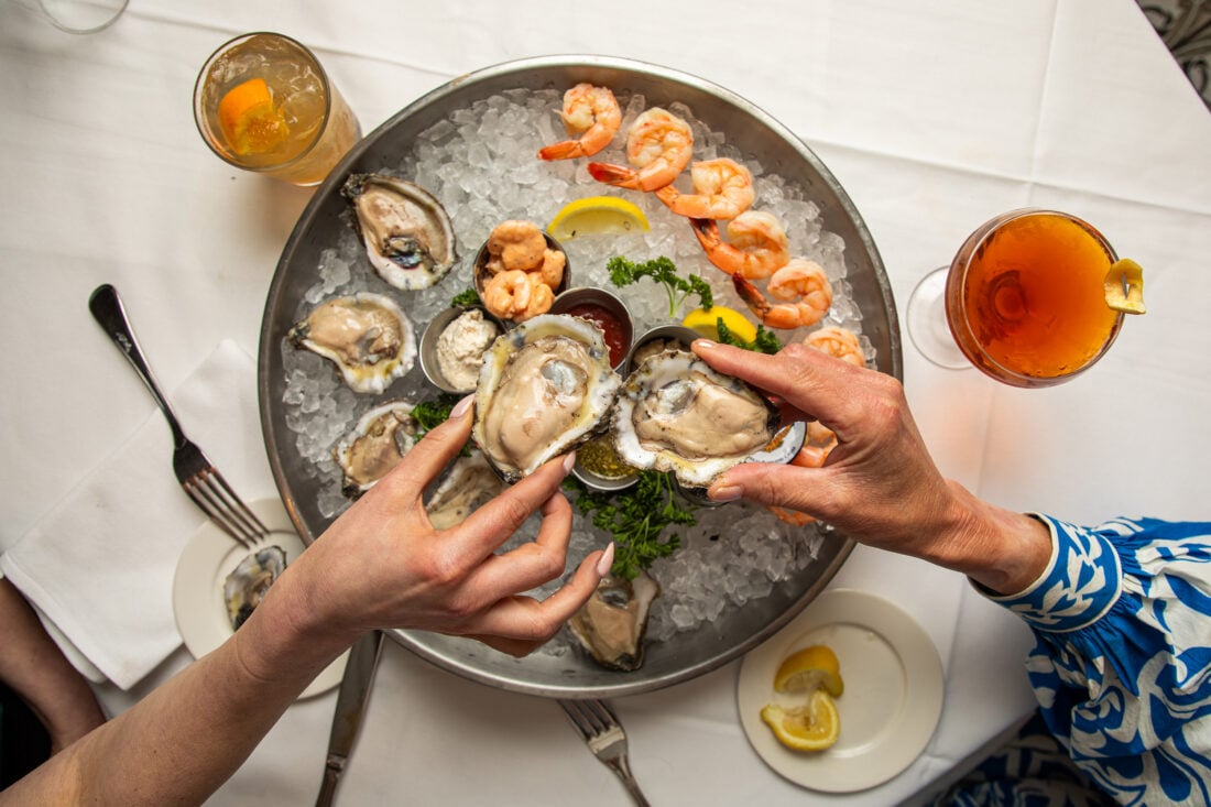 hands holding oysters above a metal tray of oysters and shrimp on ice