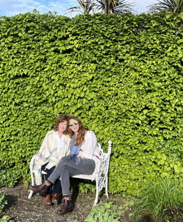 Two women sit against a green leafy wall
