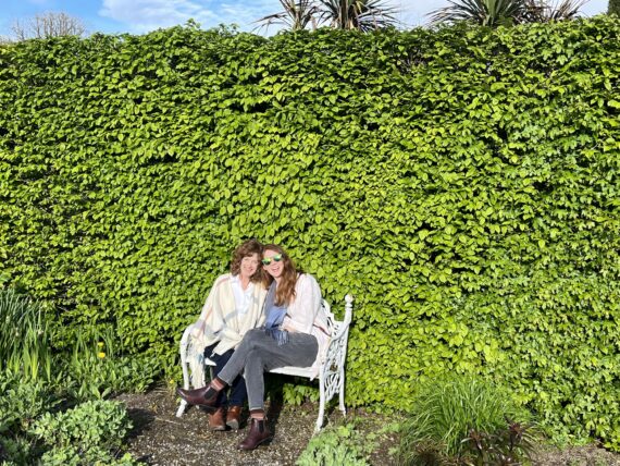 Two women sit against a green leafy wall