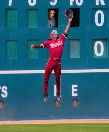 A baseball player catches a ball in mid air with a man in the scorekeeper board