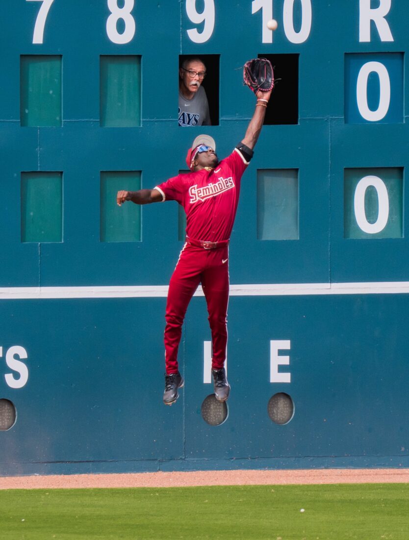 A baseball player catches a ball in mid air with a man in the scorekeeper board