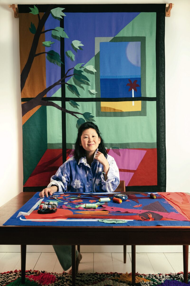 A woman in an art studio with fabric and a fabric artwork of a window behind her