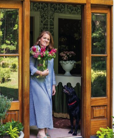 A woman stands in a home door with a black lab. She holds flowers
