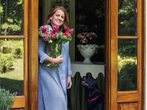 A woman stands in a home door with a black lab. She holds flowers