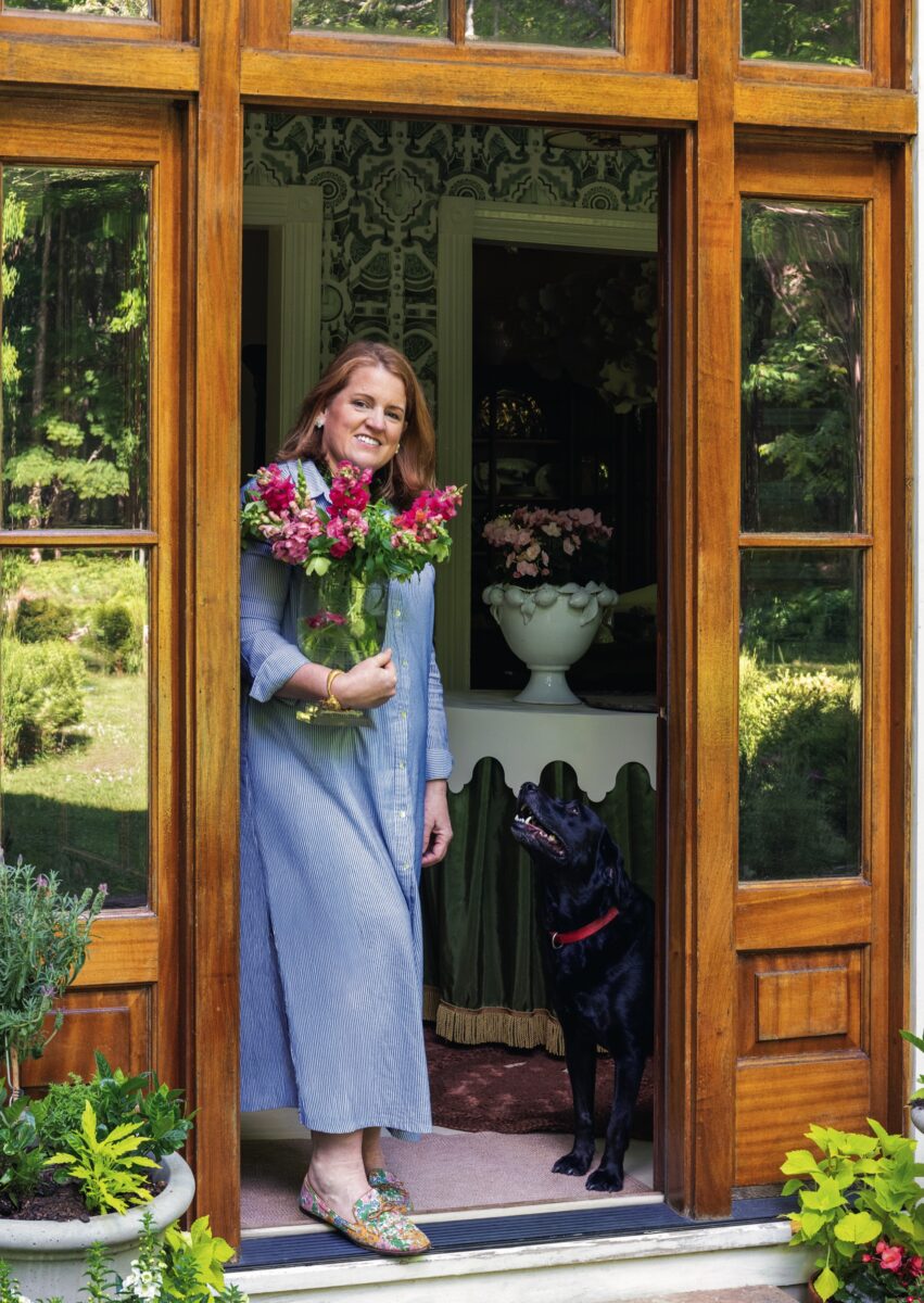 A woman stands in a home door with a black lab. She holds flowers